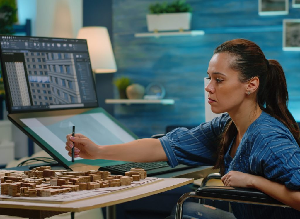 Invalid engineer looking at building model on computer designing layout and blueprint for construction. Architect in wheelchair using touch screen monitor to analyze architectural structure